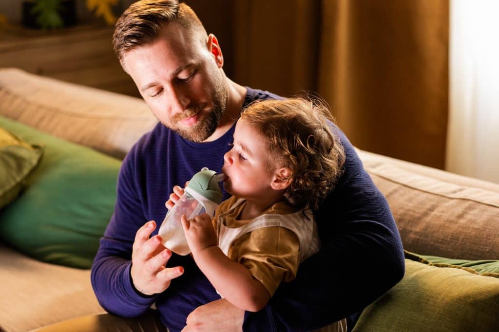 A man is holding a baby while he drinks from a bottle.